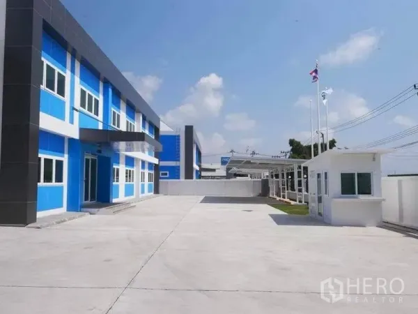 工厂 出租在Bang Bo, Samut Prakan - 厂区宽敞车道与停车区，设有门卫室与旗杆，蓝灰色办公楼立面与围墙