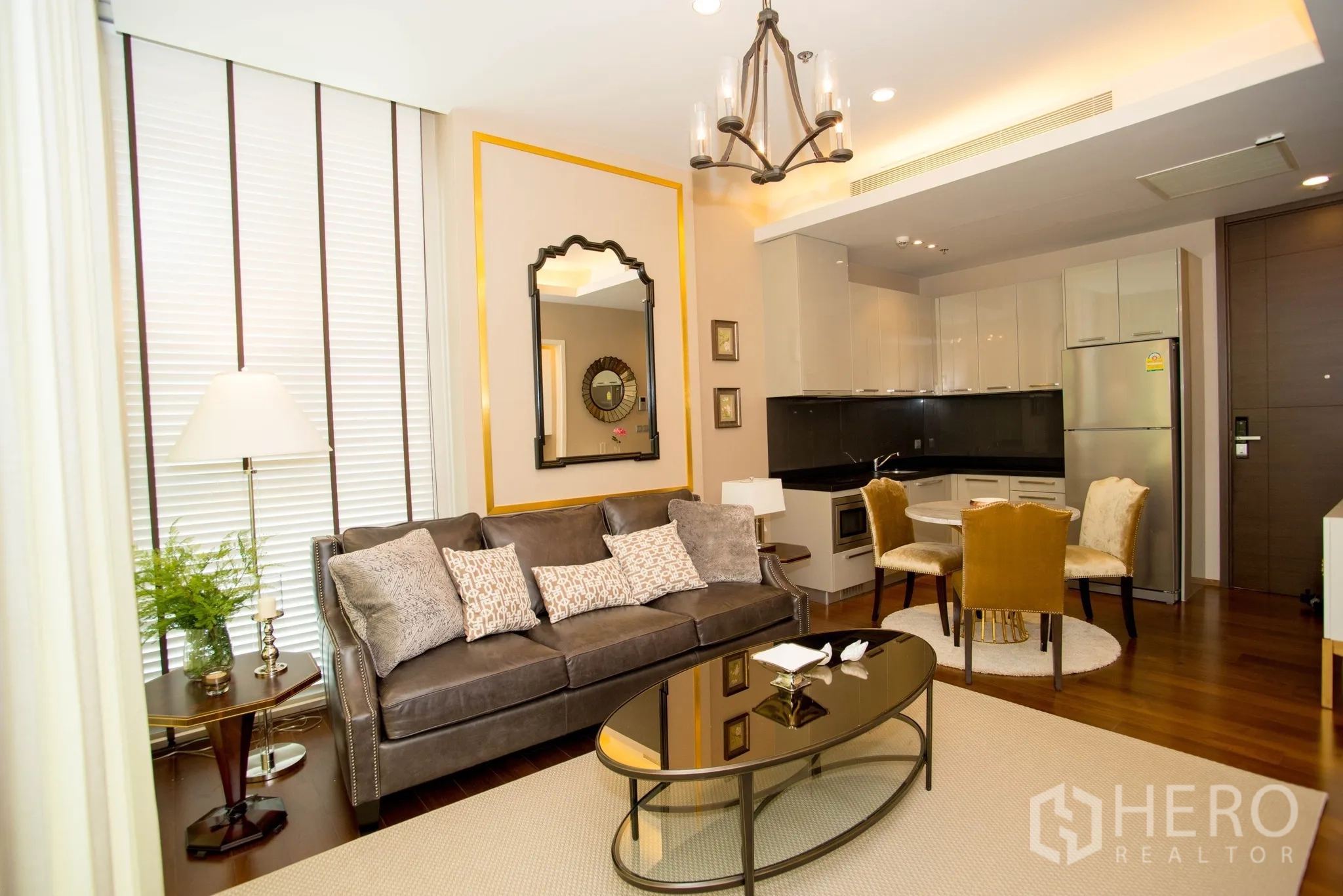 Condominium for rent in Watthana, Bangkok - Bright living room with leather sofa, chandelier, mirror feature wall, and open kitchen with white cabinets and dining set.