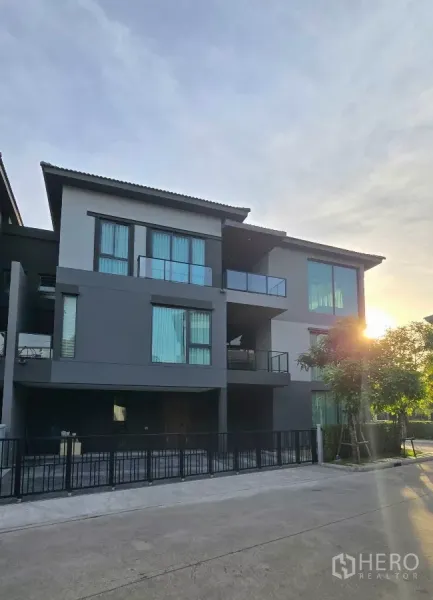 Detached House for rent in Prawet, Bangkok - Modern three-storey detached house facade with balconies and a gated driveway at sunset.
