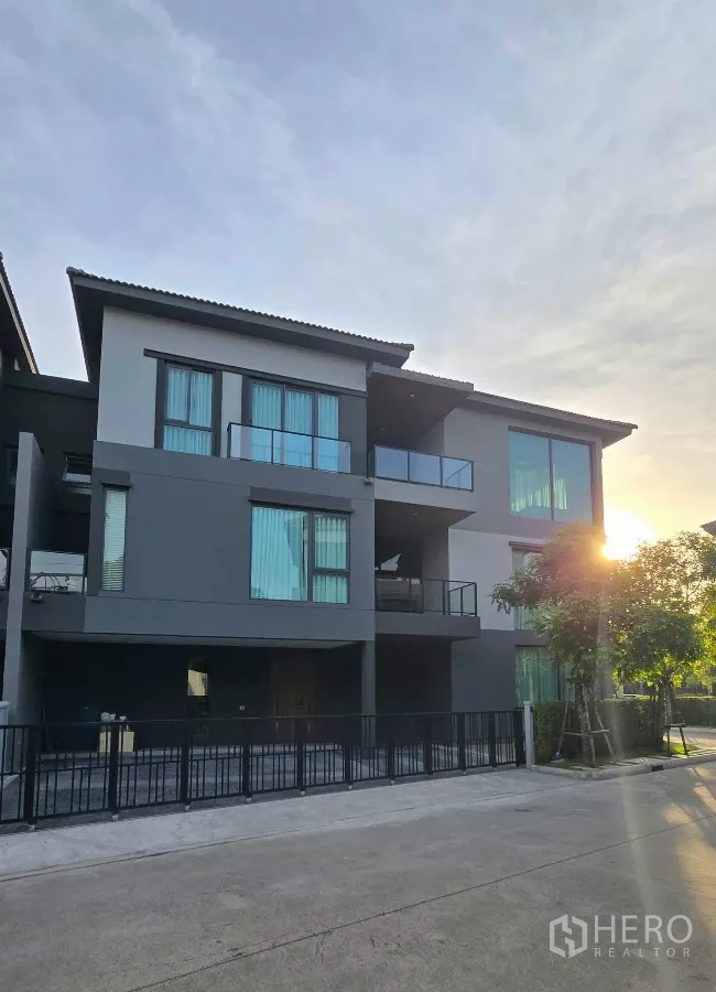 Detached House for rent in Prawet, Bangkok - Modern three-storey detached house facade with balconies and a gated driveway at sunset.