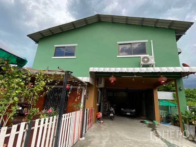 Warehouse for rent or sale in Pak Kret, Nonthaburi - Driveway leading to a covered loading bay beneath a green two-storey warehouse with plants along the fence.