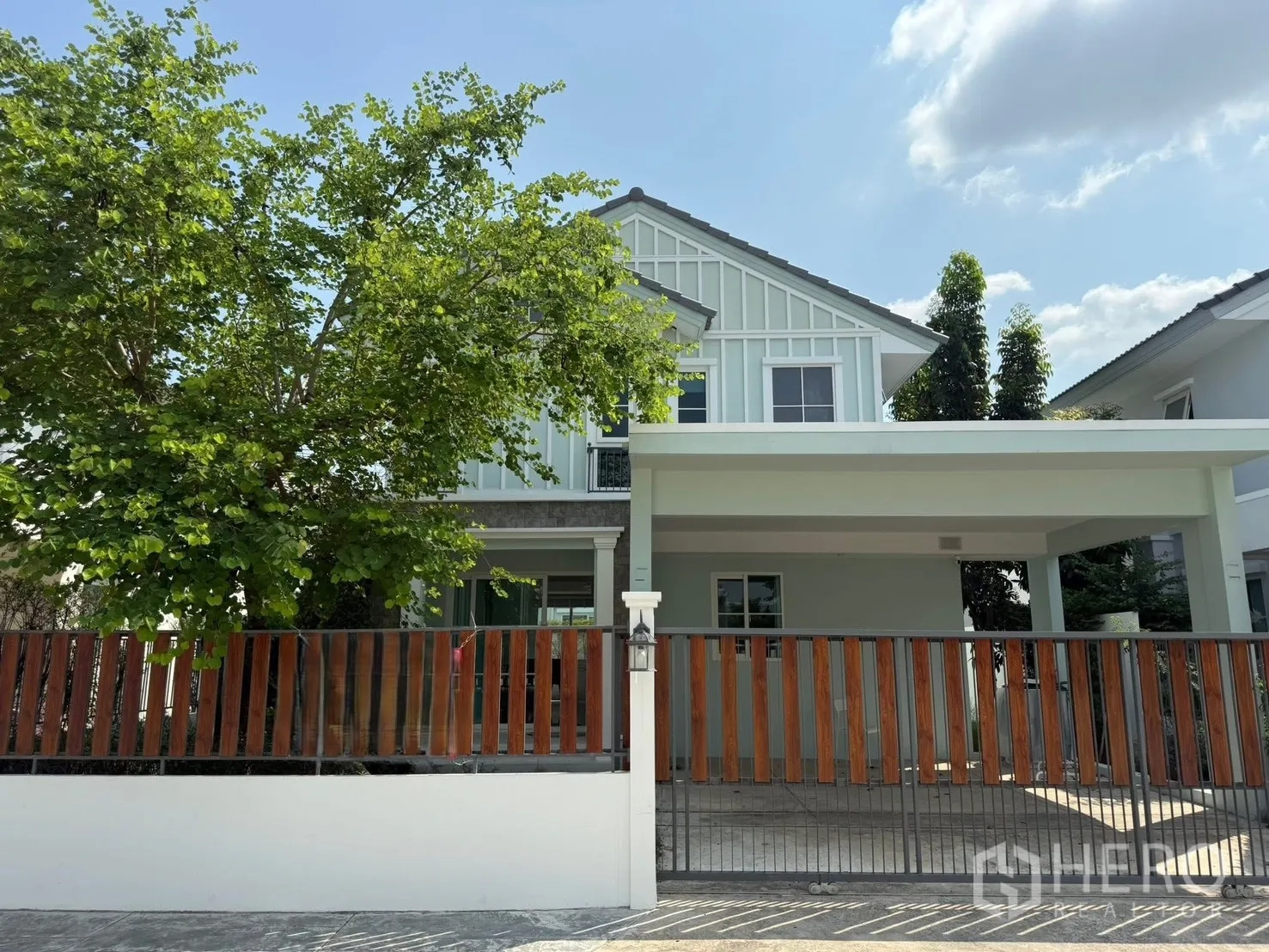 บ้านเดี่ยว for rent in Bang Phli, Samut Prakan - มุมมองด้านหน้าบ้านเดี่ยวสองชั้น มีโรงจอดรถมีหลังคา รั้วไม้ และต้นไม้ใหญ่ร่มรื่น