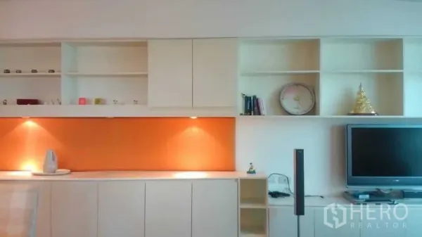 Condominium for sale in Bang Rak, Bangkok - Living room wall of built-in white cabinets with orange accent panel, open shelves and TV unit.