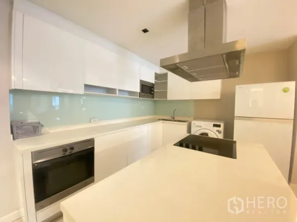 公寓 出租在Watthana, Bangkok - 明亮的U形厨房，白色橱柜，带岛台电磁炉与不锈钢烟机，玻璃挡板、冰箱和洗衣机。