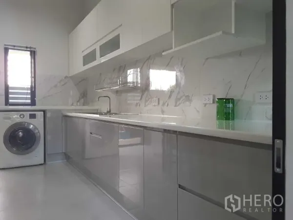 บ้าน ให้เช่าหรือขายในLam Luk Ka, Pathum Thani - ห้องครัวโทนขาวสว่าง ผนังลายหินอ่อน เคาน์เตอร์ยาว ตู้ลอย และเครื่องซักผ้าข้างหน้าต่าง