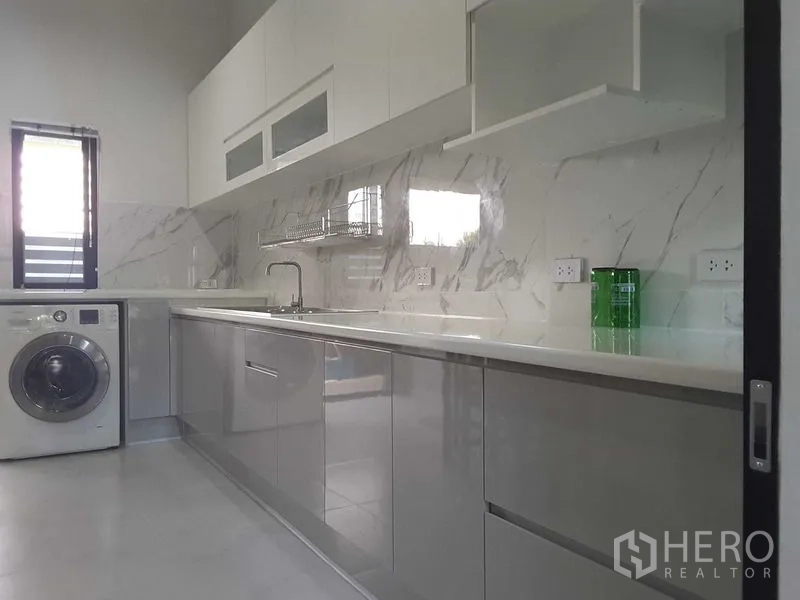 บ้าน ให้เช่าหรือขายในLam Luk Ka, Pathum Thani - ห้องครัวโทนขาวสว่าง ผนังลายหินอ่อน เคาน์เตอร์ยาว ตู้ลอย และเครื่องซักผ้าข้างหน้าต่าง