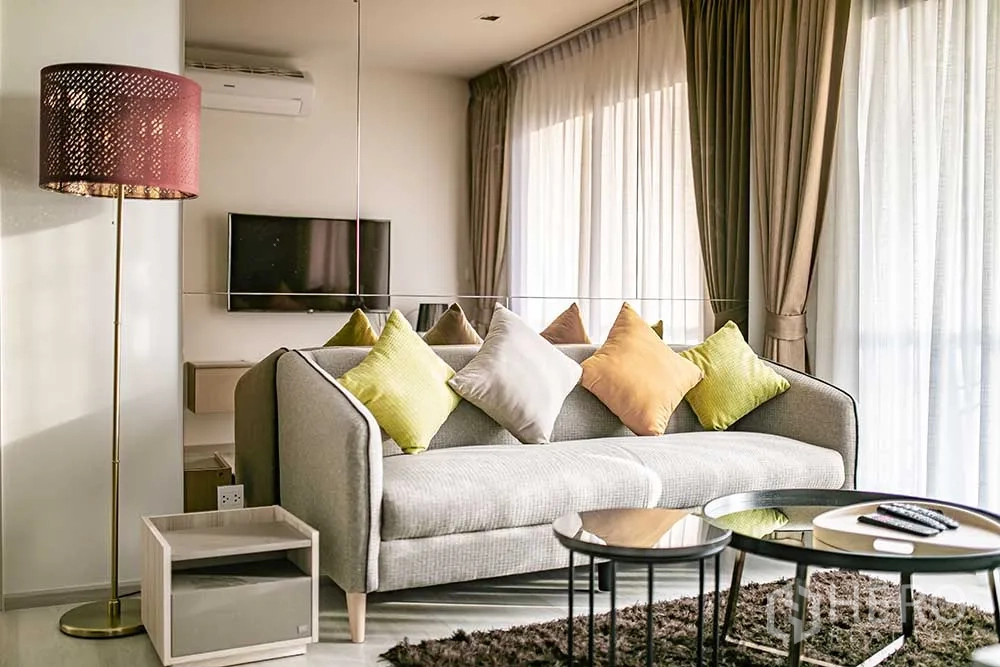 Condominium for rent in Pathum Wan, Bangkok - Bright living room with grey sofa, colorful cushions, wall-mounted TV, round coffee tables, floor lamp, and large curtained windows.