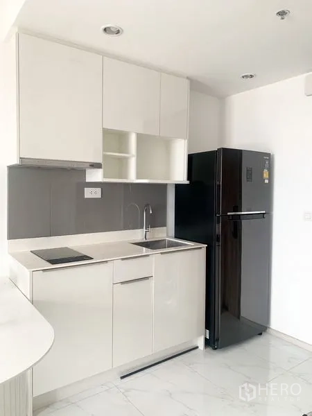 Condominium for sale in Bang Na, Bangkok - Compact white kitchenette with electric hob, sink, cabinets, and a black refrigerator.