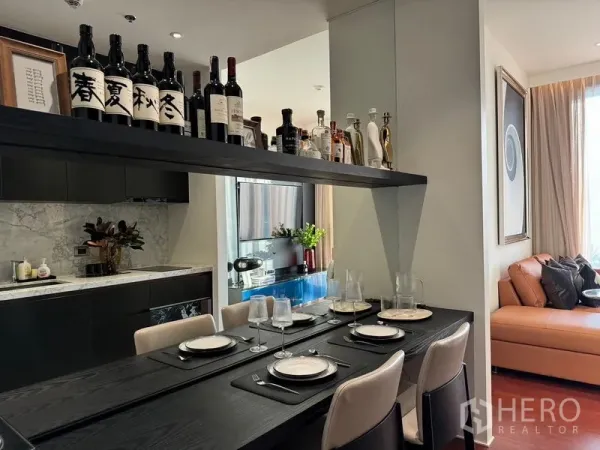 Condominium for sale in Watthana, Bangkok - Open-plan dining and kitchen with marble backsplash, black cabinetry, a table set for four and a display shelf of bottles.