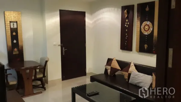 Condominium for rent or sale in Khlong Toei, Bangkok - Living room with leather sofa, glass coffee table, dark wood door, wall art, and small dining set.