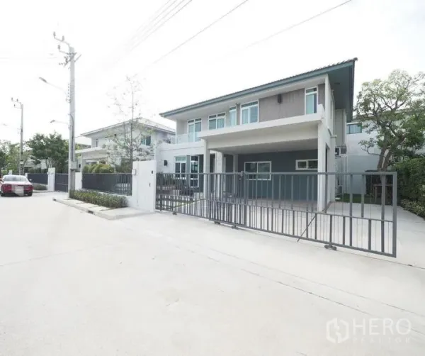 Detached House for rent in Prawet, Bangkok - Angled exterior view showing wide driveway and sliding metal gate on a quiet street.