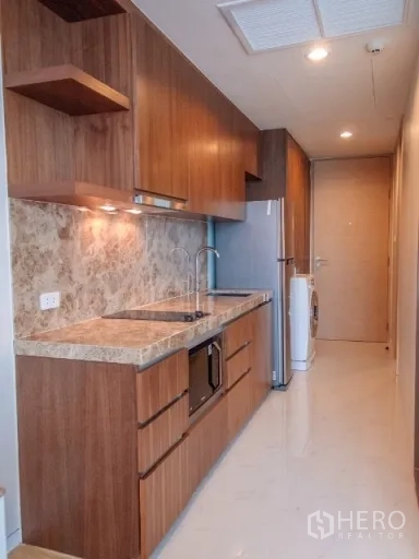 Condominium for rent in Watthana, Bangkok - Galley kitchen with warm wood cabinetry, stone countertops, electric cooktop, microwave, fridge, and washer.