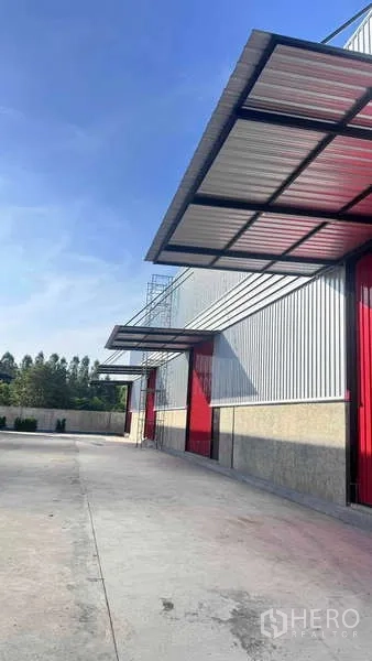 货栈 出租在Khlong Sam Wa, Bangkok - 仓库立面装卸雨棚与红色卷帘门，前方为大面积硬化场地。