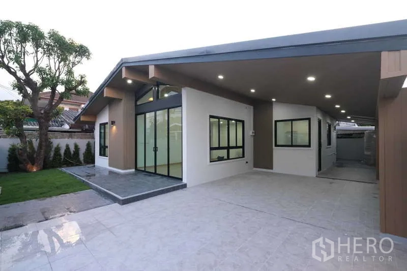 独立式住宅 出售在Bueng Kum, Bangkok - 现代单层独栋别墅外观，带遮棚车位、大窗户、铺砖车道与小草坪，傍晚灯光柔和