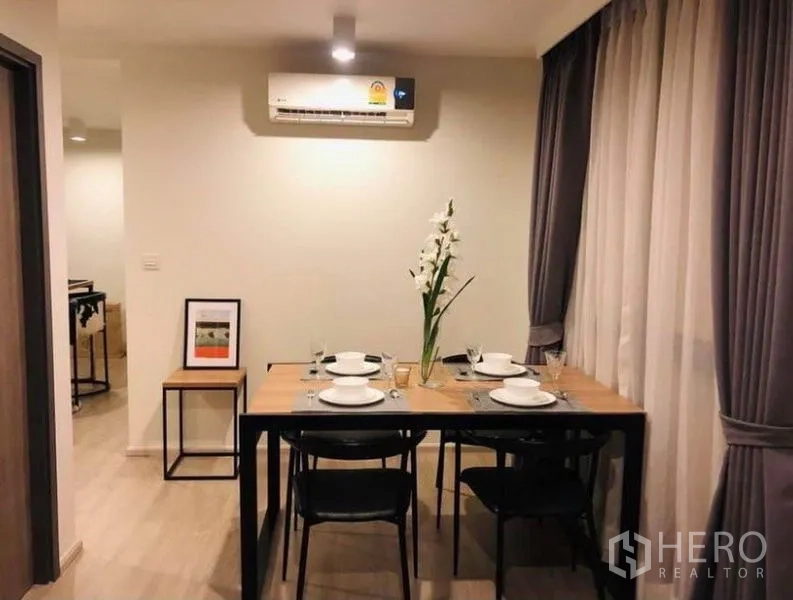 Condominium for rent or sale in Pathum Wan, Bangkok - Dining area with a wooden table set for four beneath an air conditioner and sheer drapes.