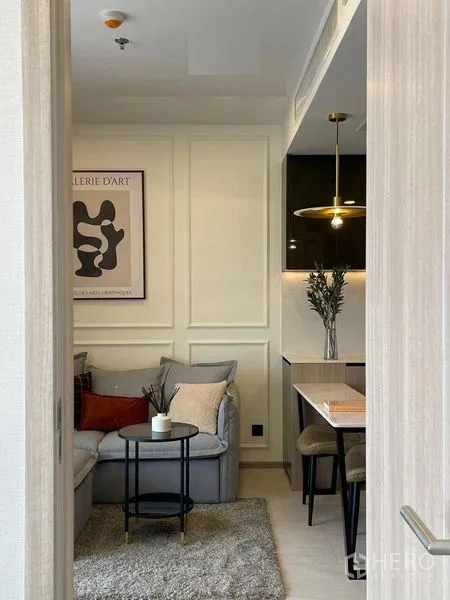 公寓 出租在Pathum Wan, Bangkok - 温馨客厅，L型沙发与装饰画，旁边是餐区和备餐台。