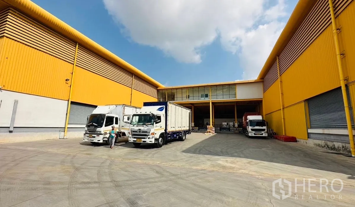 Warehouse for rent in Lat Krabang, Bangkok - Truck yard between warehouse buildings with multiple loading bays, roller shutter doors, and parked delivery trucks.
