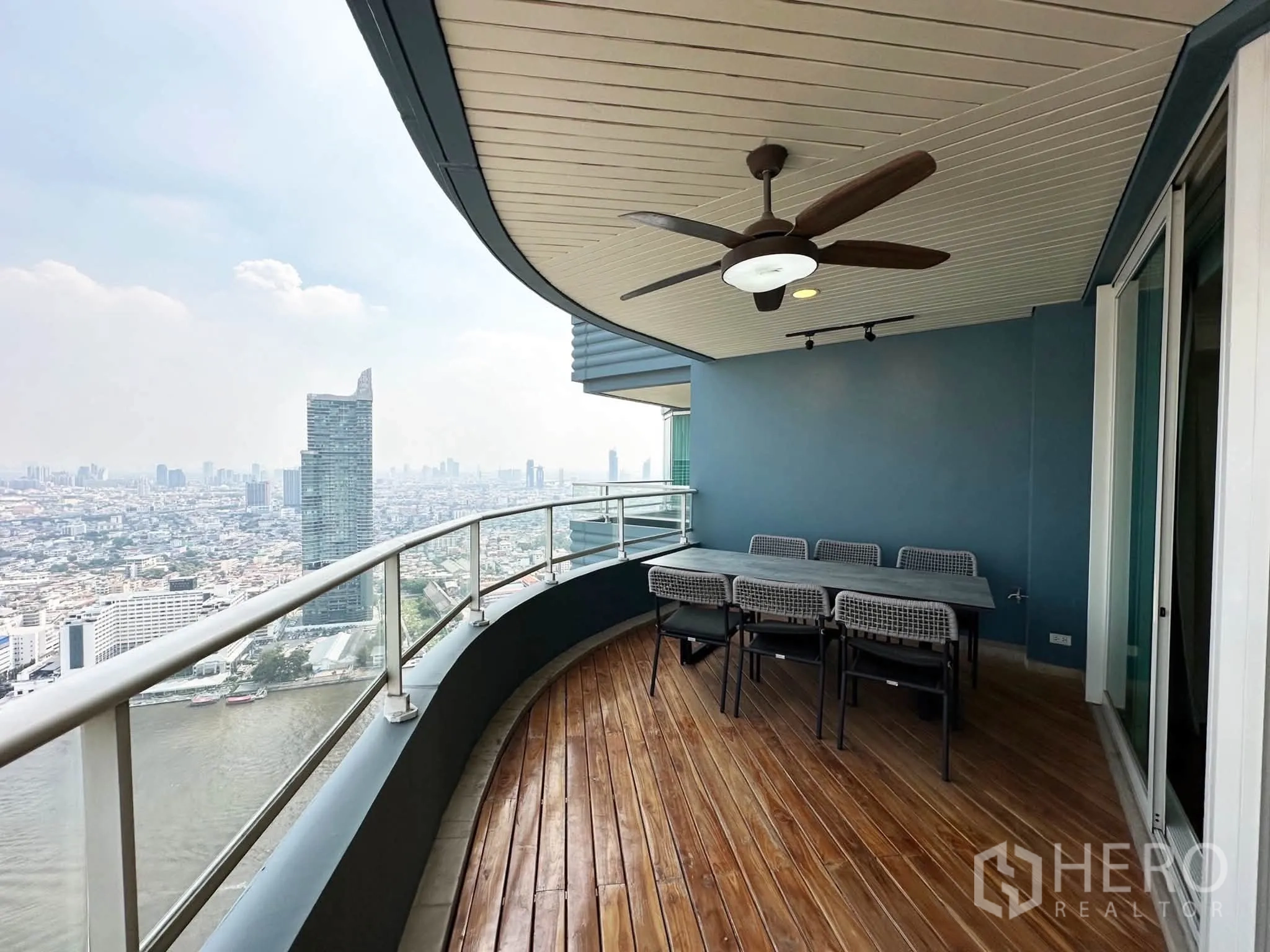 Condominium for sale in Khlong San, Bangkok - Curved balcony with wooden decking, ceiling fan, and outdoor dining set overlooking the river and skyline.