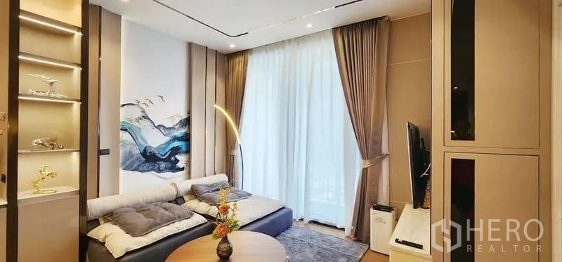 คอนโด ให้เช่า - ห้องนั่งเล่นอบอุ่น โซฟา ผนังทีวี และผ้าม่านโปร่งสู่ระเบียง