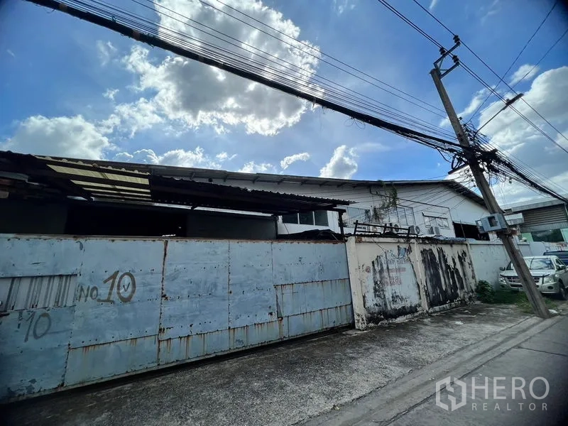 Warehouse for rent in Suan Luang, Bangkok - Street view of gated industrial entrance with tall metal sliding gate and overhead power lines.