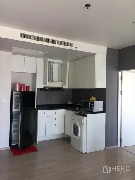 Condominium for rent in Watthana, Bangkok - Compact corner kitchen with white cabinets, black countertops, refrigerator, microwave, washing machine, and light wood floors.