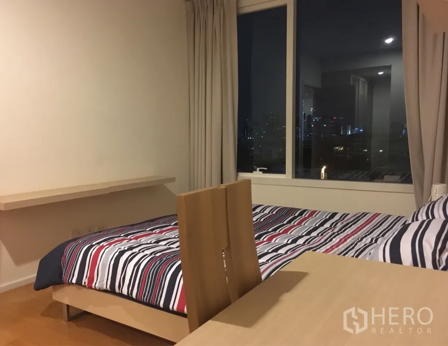 คอนโด ให้เช่าในWatthana, Bangkok - ห้องนอนผ้าปูที่นอนลายทาง มีโต๊ะทำงานและหน้าต่างบานใหญ่เห็นวิวเมืองยามค่ำ