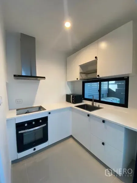 Townhouse for rent in Saphan Sung, Bangkok - Compact L-shaped white kitchen with oven, cooktop, extractor hood and window for ventilation.
