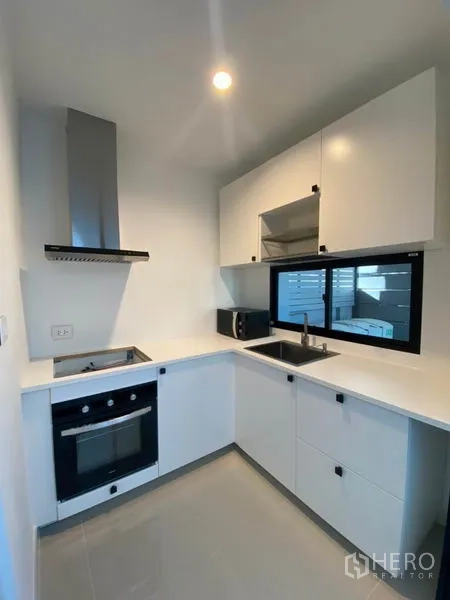 Townhouse for rent in Saphan Sung, Bangkok - Compact L-shaped white kitchen with oven, cooktop, extractor hood and window for ventilation.