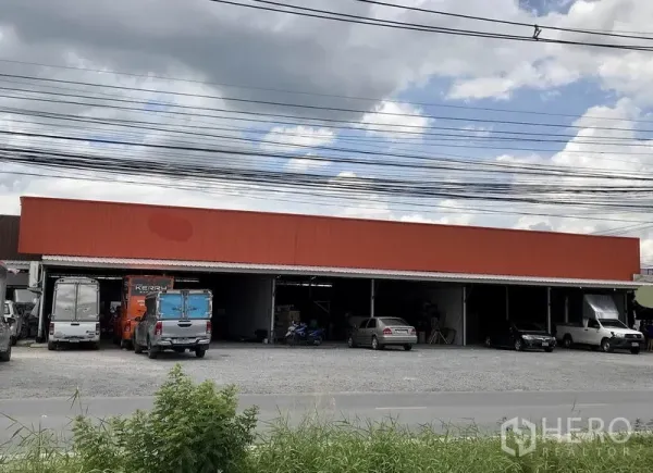 货栈 出租在Pathum Thani - 单层橙色立面的仓库外观，装卸口敞开，前方碎石场地宽阔，停有卡车与小车。