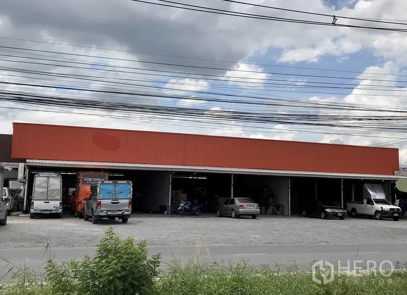 货栈 出租在Pathum Thani - 单层橙色立面的仓库外观，装卸口敞开，前方碎石场地宽阔，停有卡车与小车。