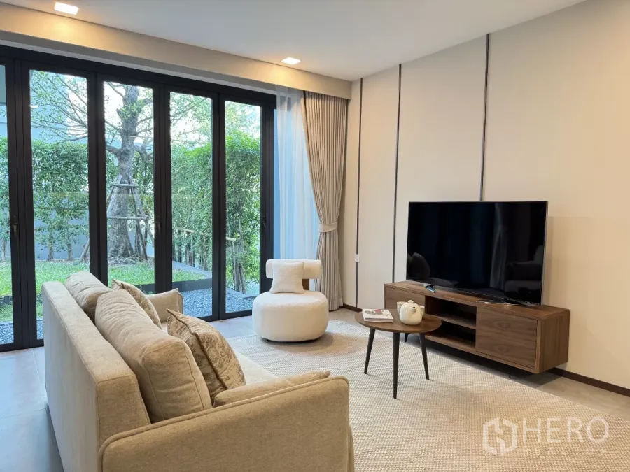 Detached House for rent in Saphan Sung, Bangkok - Bright living room with beige sofa, TV console and floor-to-ceiling glass doors opening to a green garden.