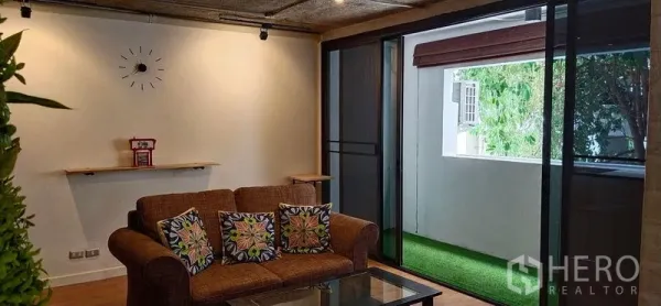 公寓 出售在Watthana, Bangkok - 客厅棕色沙发与玻璃茶几，推门通往人造草坪阳台。