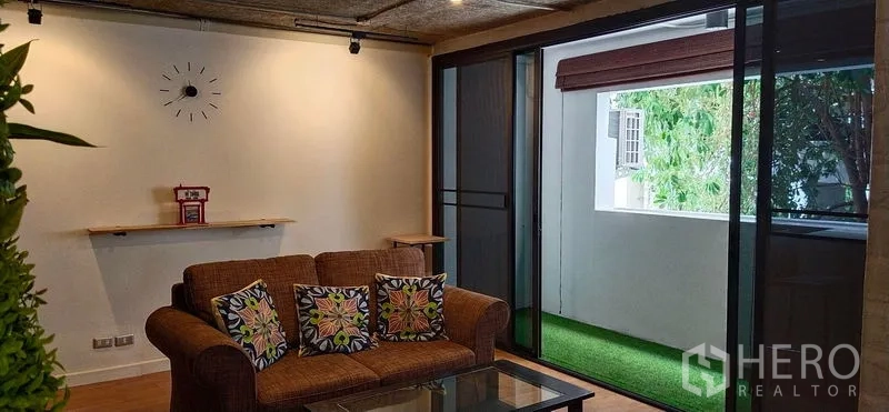 公寓 出售在Watthana, Bangkok - 客厅棕色沙发与玻璃茶几，推门通往人造草坪阳台。