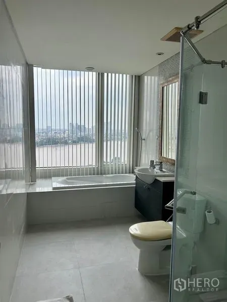 คอนโด ให้เช่า - ห้องน้ำสมัยใหม่พร้อมอ่างอาบน้ำ กั้นอาบน้ำกระจก และวิวเมืองกับแม่น้ำผ่านหน้าต่างสูง