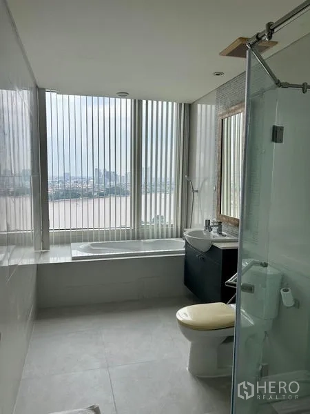 คอนโด ให้เช่า - ห้องน้ำสมัยใหม่พร้อมอ่างอาบน้ำ กั้นอาบน้ำกระจก และวิวเมืองกับแม่น้ำผ่านหน้าต่างสูง
