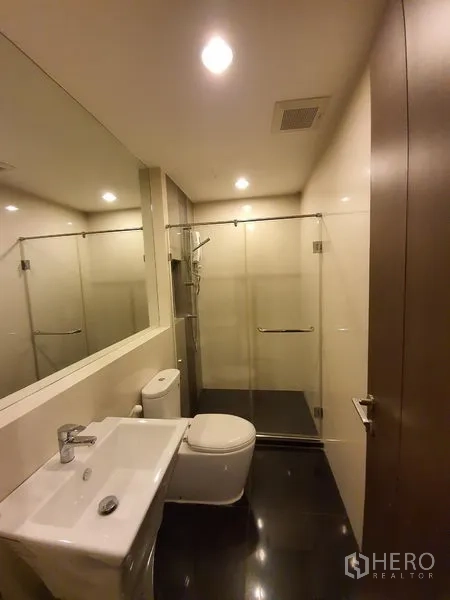Condominium for rent in Sathon, Bangkok - Modern bathroom with large mirror, glass-enclosed shower and white basin and toilet.