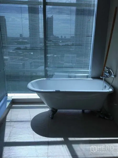 Condominium for sale in Bang Rak, Bangkok - Bathroom with freestanding clawfoot bathtub beside a large window with blinds and skyline view.