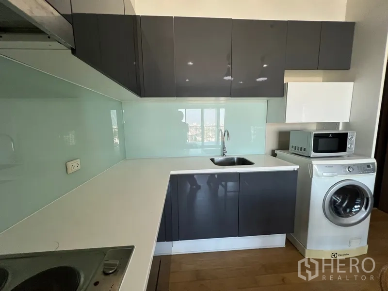 Condominium for rent or sale in Watthana, Bangkok - Compact L-shaped kitchen with sink, upper cabinets, microwave and front-load washer