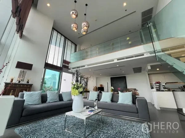 Condominium for sale in Khlong San, Bangkok - Double-height living room with mezzanine, pendant lights, gray sofas, and a glass staircase beside an open kitchen.