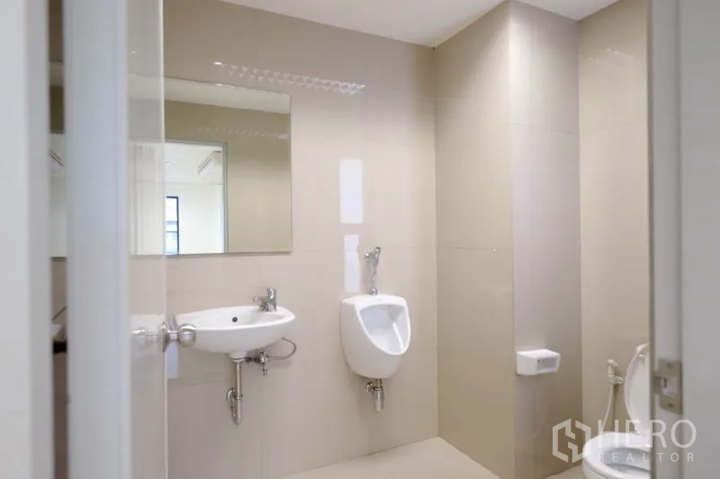 办公空间 出租在Chatuchak, Bangkok - 明亮的办公室卫生间，设有洗手盆、小便器和大镜子，米色瓷砖墙面。