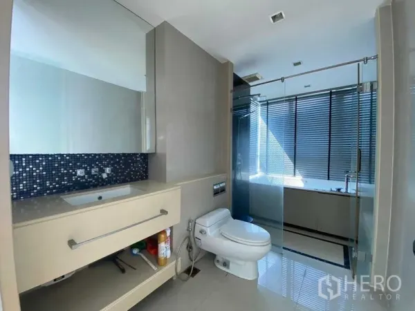 Condominium for sale in Ratchathewi, Bangkok - Modern bathroom with wide vanity, toilet and glass-enclosed shower and tub beside window blinds.