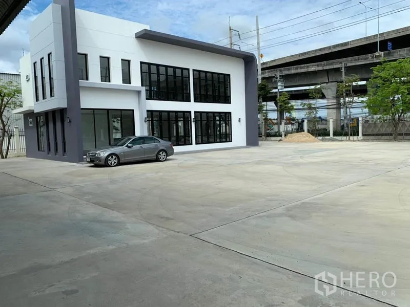 Warehouse for rent in Bang Phli, Samut Prakan - Two-storey glass-front office beside a large parking and loading yard with an elevated roadway in the background.