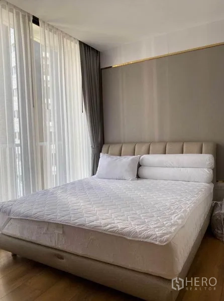 Condominium for rent in Khlong Toei, Bangkok - Sunlit master bedroom with upholstered bed, neutral tones, and full-height curtains.