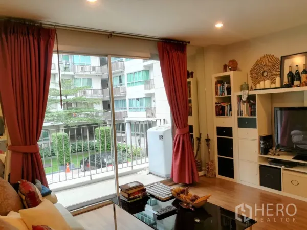 Condominium for rent in Watthana, Bangkok - TV wall with built-in shelving and large windows dressed with red curtains opening to a balcony.