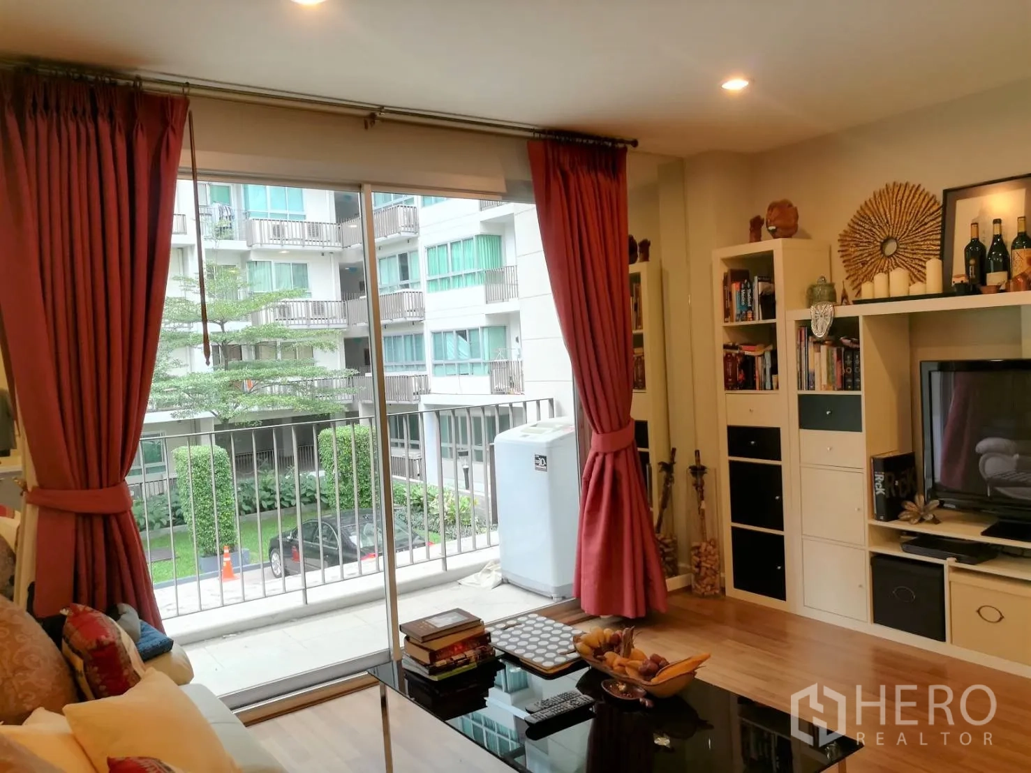 Condominium for rent in Watthana, Bangkok - TV wall with built-in shelving and large windows dressed with red curtains opening to a balcony.