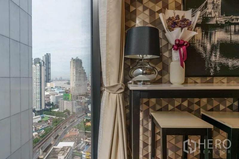 คอนโด ให้เช่าหรือขายในBang Rak, Bangkok - มุมนั่งข้างหน้าต่างพร้อมโคมไฟและภาพตกแต่ง มองเห็นตึกสูงและรางรถไฟฟ้า BTS