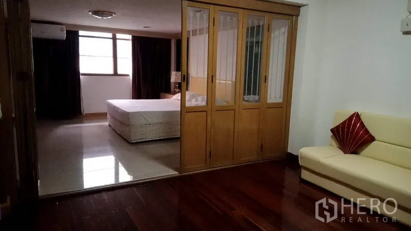 Townhouse for rent in Khlong Toei, Bangkok - Large suite with glass‑panel wardrobe partition, cream sofa and polished wood flooring.