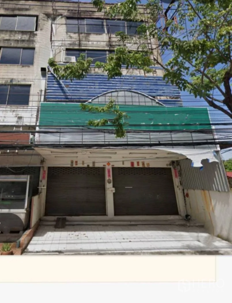 Shophouse for rent in Bang Na, Bangkok - Four-storey shophouse exterior with double roll-up shutters and a small tiled forecourt shaded by a tree.