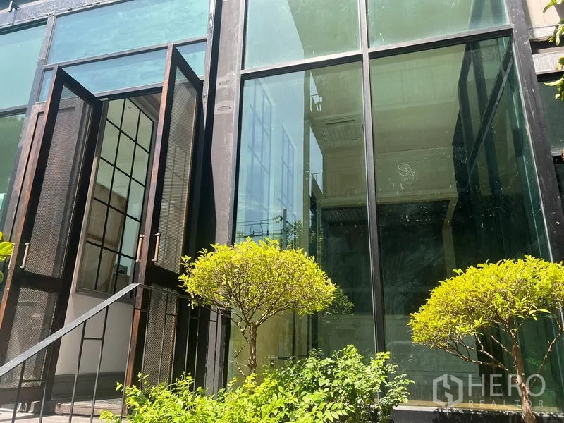 零售空间 出租在Khlong Toei, Bangkok - 临街玻璃店面，黑色钢框双开门敞开，入口处修剪灌木，通罗商圈零售单位外观