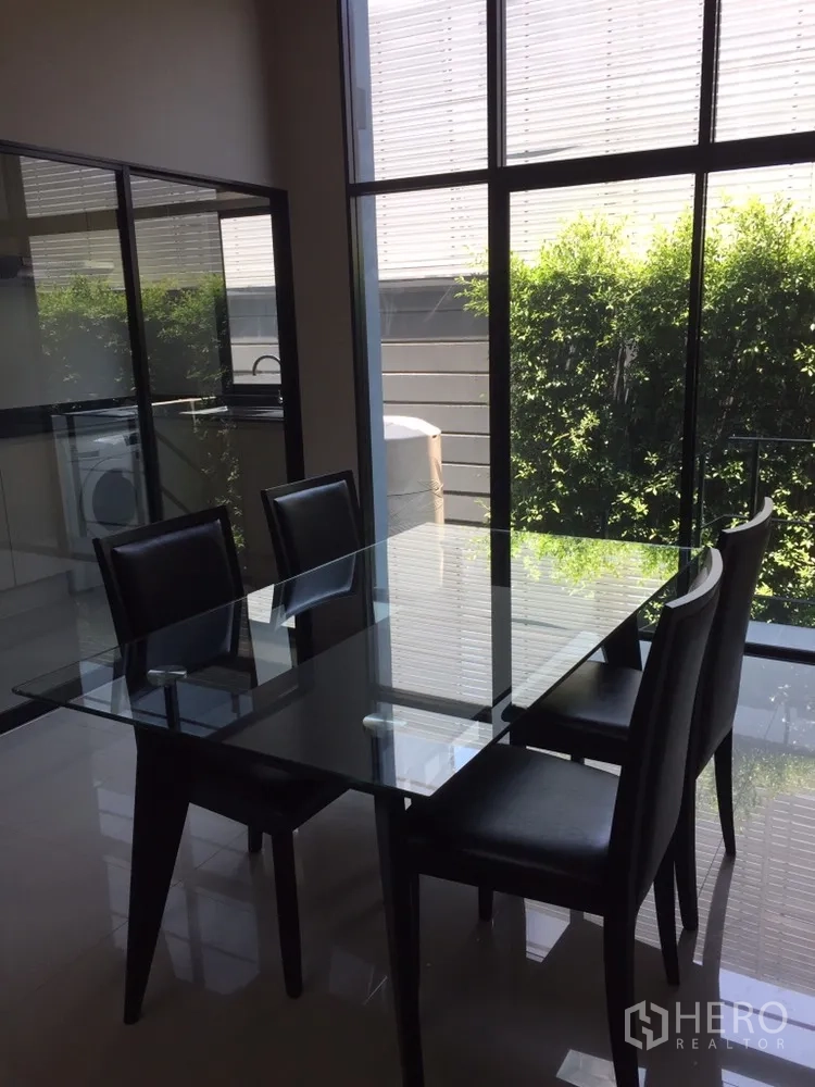 Townhouse for rent in Suan Luang, Bangkok - Dining space with a glass table and four chairs by floor-to-ceiling windows facing greenery.